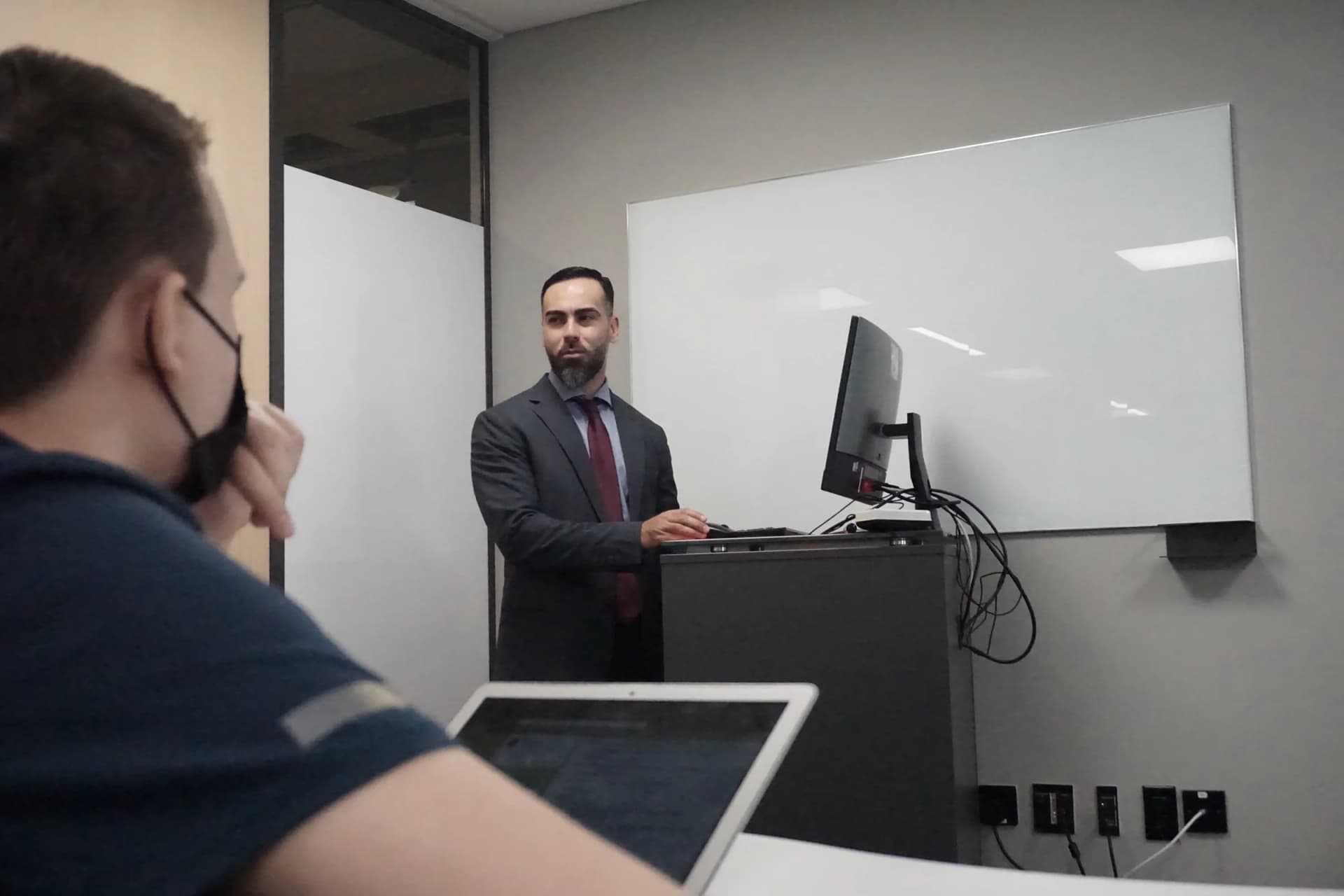 Dr. Alex Jones teaching from a lectern beside a whiteboard, a student in the foreground taking notes